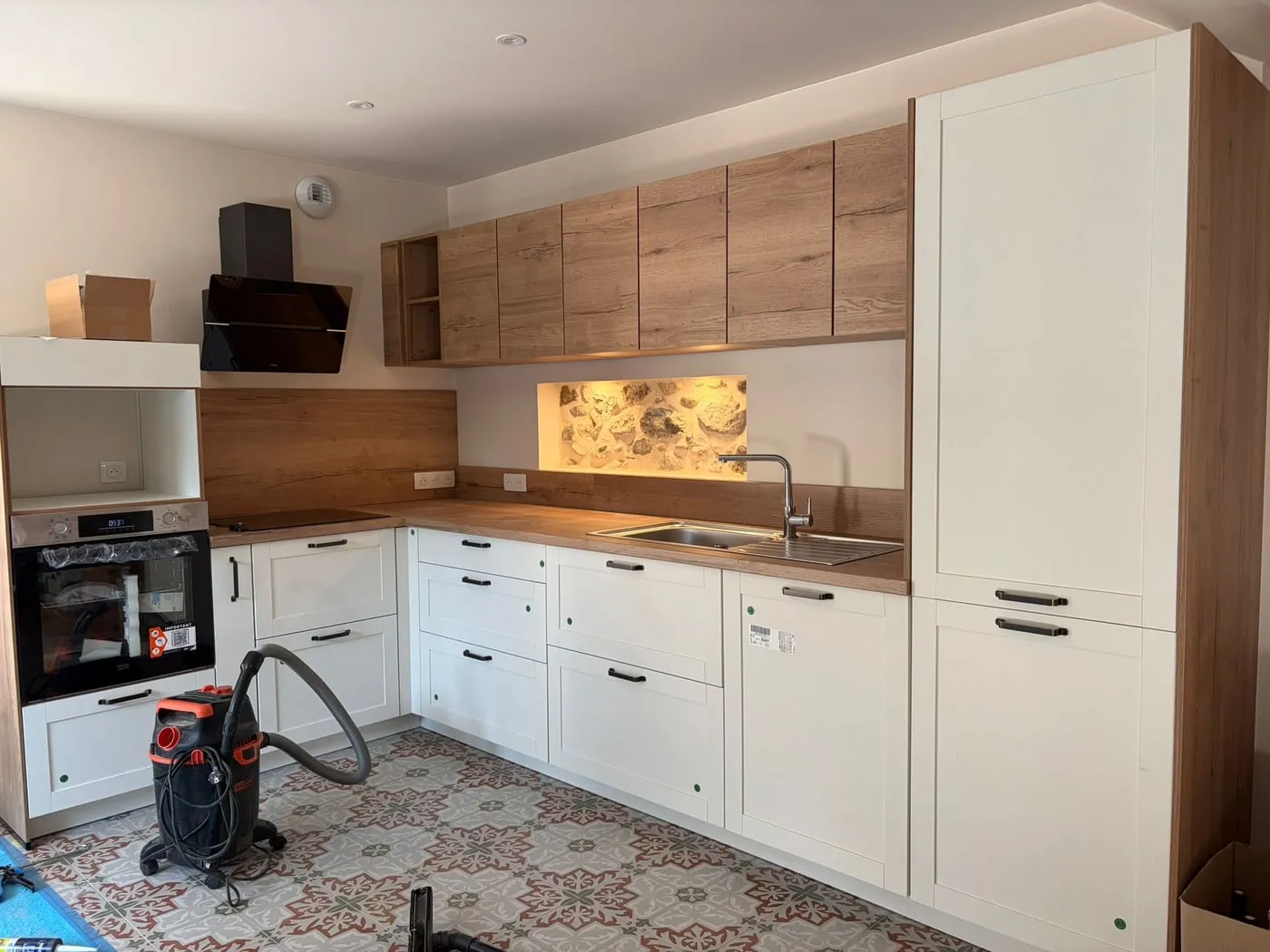 La même pièce après pose : cuisine sur mesure en bois et façades blanches, niche en pierre mise en lumière